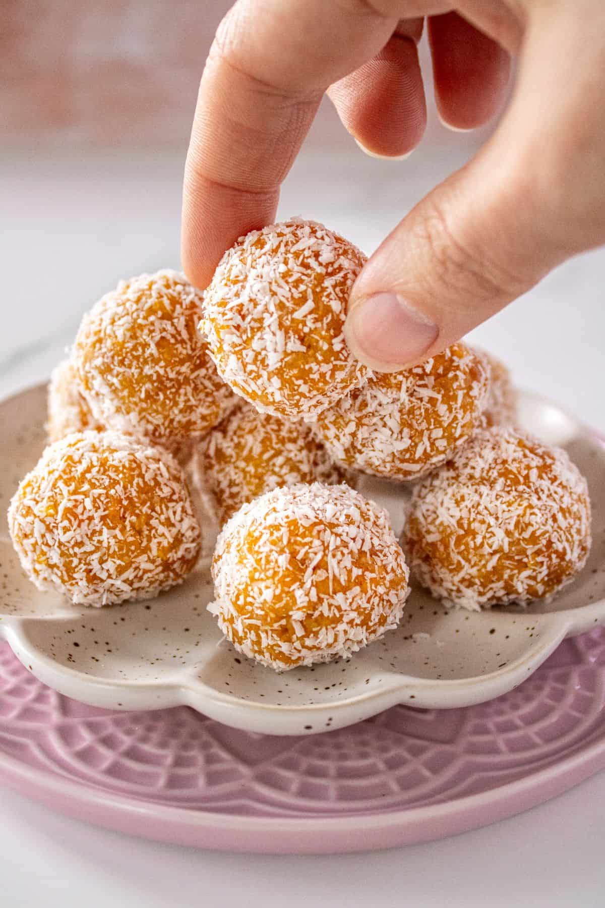 Hand reaching in to pick up a Peach Bliss Ball, from a white plate which is sitting on a pink plate.