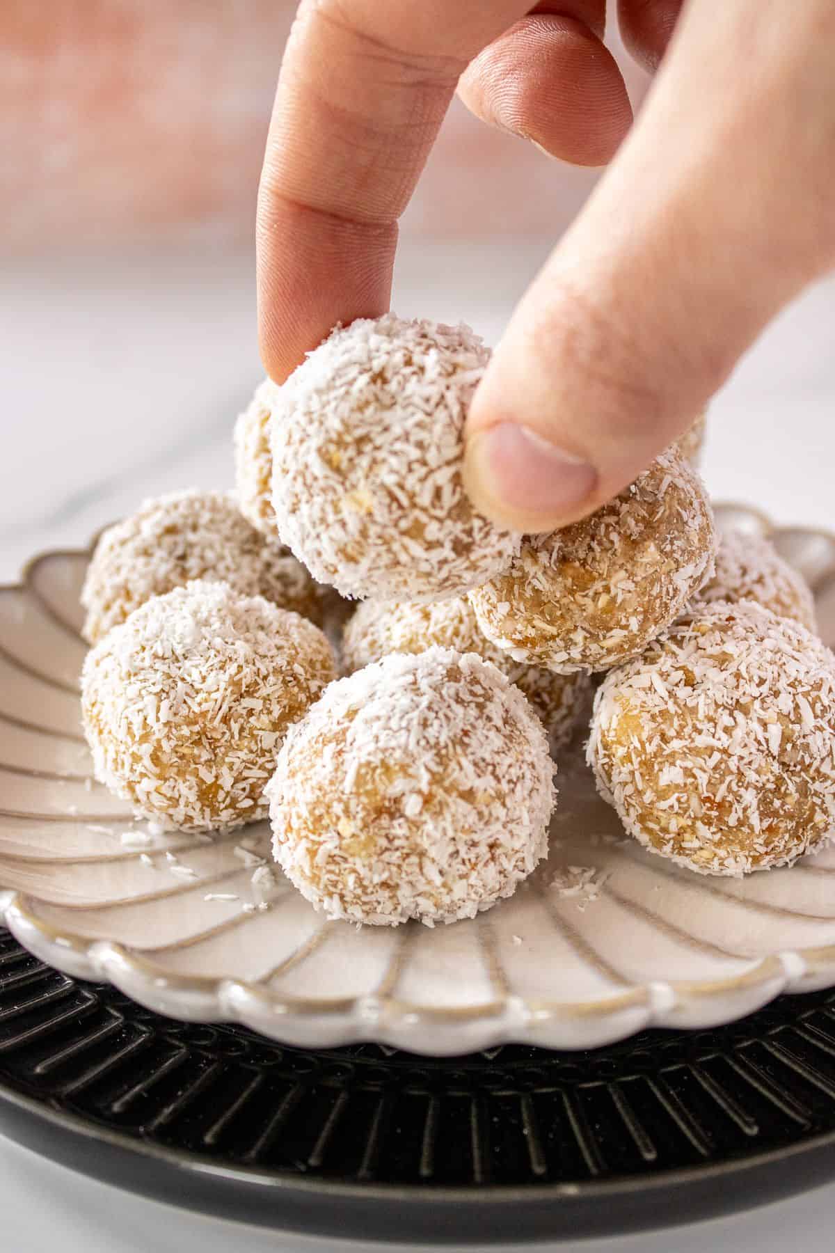 Hand reaching in to pick up a Pear Bliss Ball, from a white plate which is sitting on a black plate.